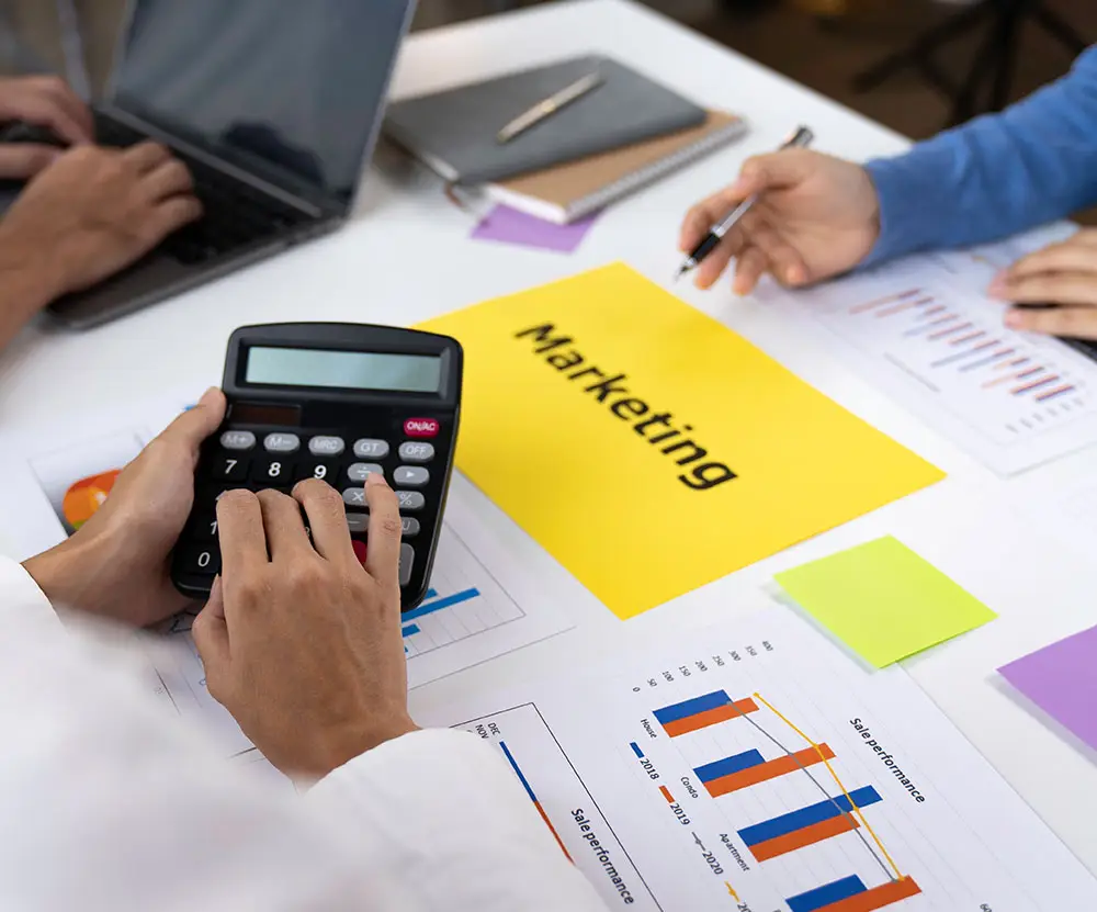 Person using a calculator during a marketing meeting, surrounded by laptops and documents.