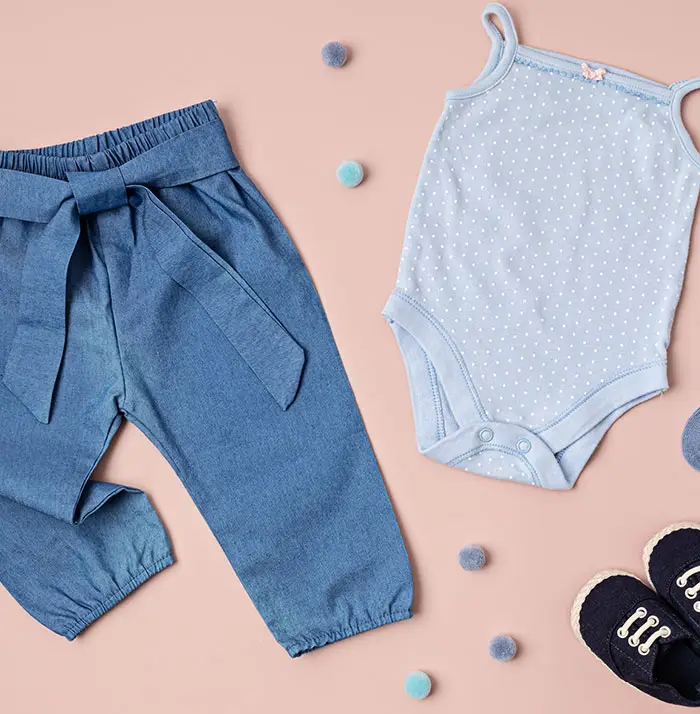 Baby outfit displayed on a soft pink background, including blue trousers with a tie, a polka dot bodysuit, socks, and tiny black shoes.