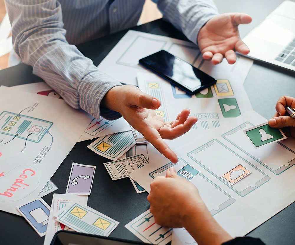 Designers discussing mobile app interface cards on a table, indicating user interface planning and teamwork.