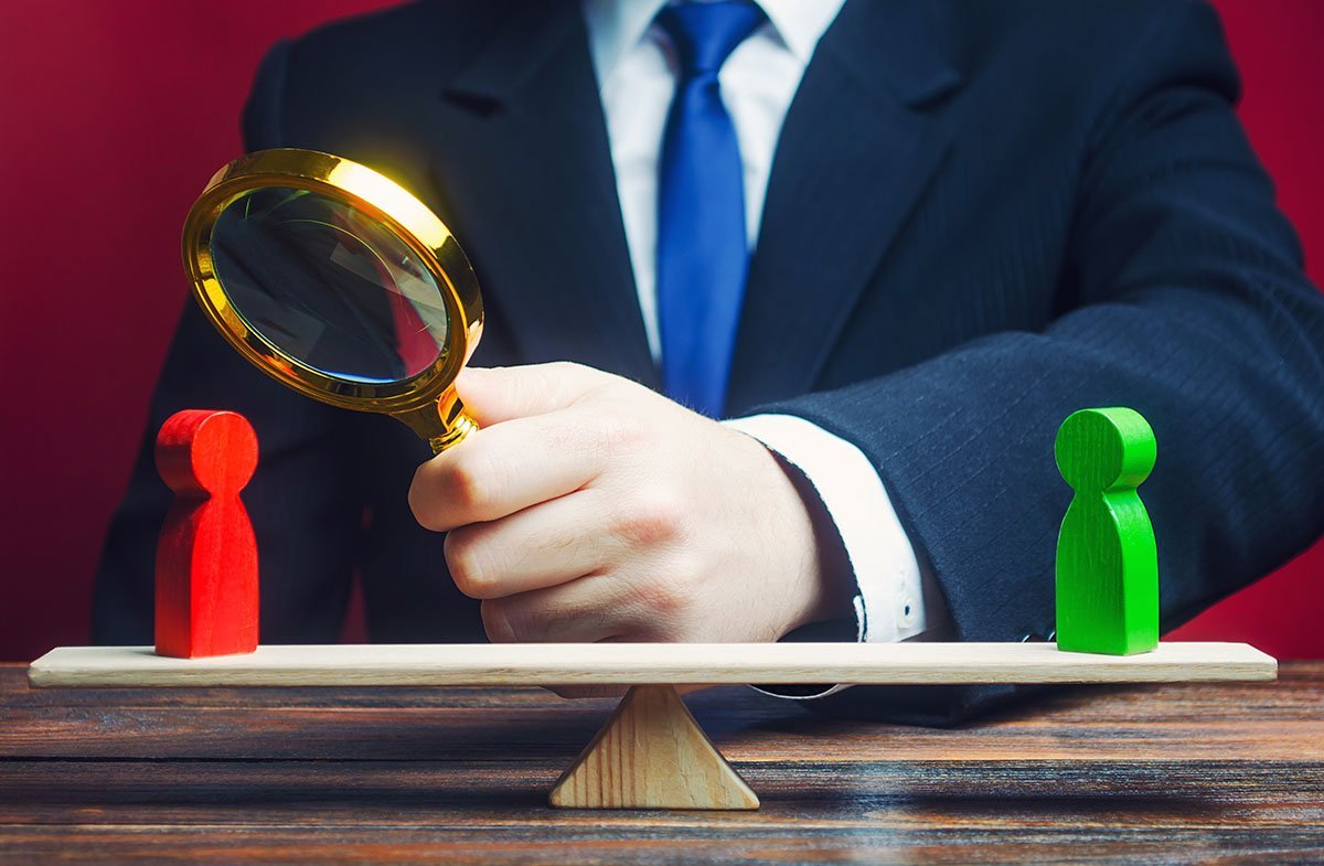 A businessperson examines two human figures on a seesaw with a magnifying glass, symbolizing scrutiny or evaluation.