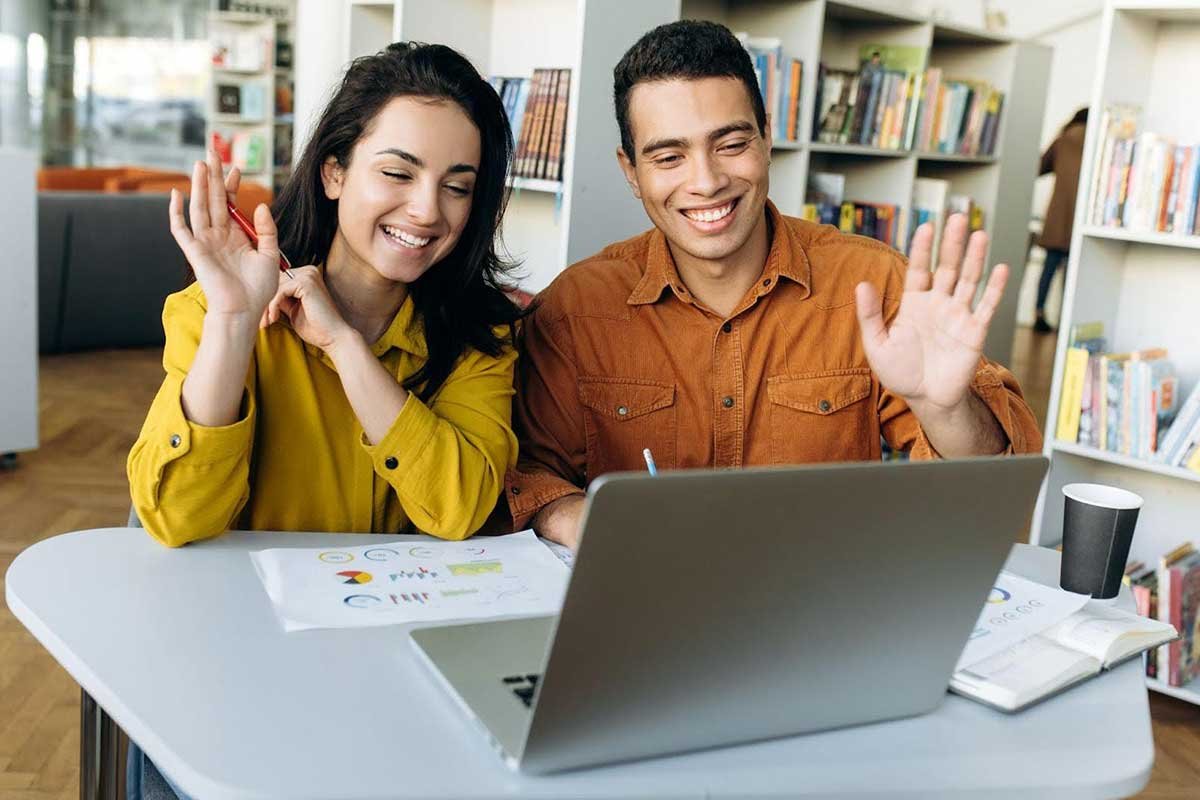 Two young professionals happily waving at a laptop screen during a video call in a modern office setting.